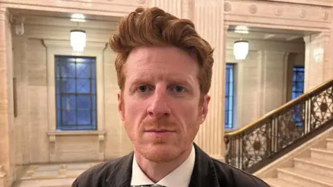 O'Toole has ginger long hair in a quiff. It is a head and shoulders shot and he is looking directly at the camera.