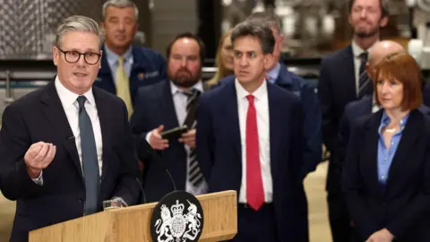 PA Media Sir Keir Starmer makes a speech in front of a wooden podium. He has his hand clenched. He is wearing a suit and navy tie. Rachel Reeves, in a blue shirt and suit jacket, and Ed Miliband, in a red tie and white shirt and navy suit, look on during a tour of a factory in Cheshire.