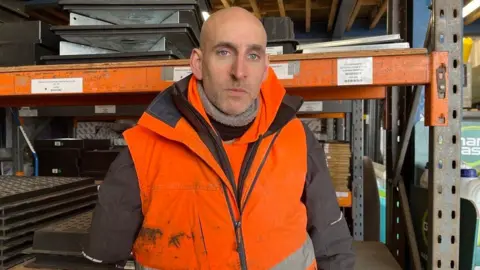 A bald man with stubble looking unhappy. He is wearing a fluorescent orange jacket and has one arm. He is standing in front of shelves in a construction unit.