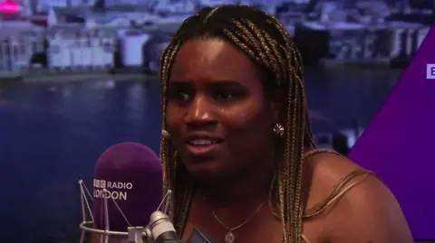 BBC Esther Obigbesan speaks into a BBC Radio London microphone in a studio, with the Thames and London skyline visible on the backdrop behind her.
