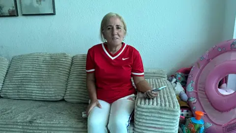 BBC is an older woman in Nike Red Football shirt and Adidas White Sitting Sitting on a cold sofa in an apartment with her cell phone
