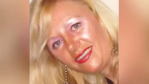 Tina has blond long wavy hair and blue eyes. She is wearing pink eye shadow and dark eyeliner. She has red lipstick on and long silver dangly earrings. 