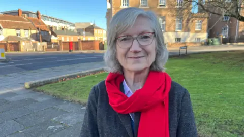 Councillor Heather Shearer is smiling at the camera. She is in a village setting, she is wearing pale rimmed glasses, a grey jacket and a red scarf. 