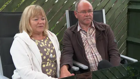 This is an image of Helen and Mark Saltern from 2020. They are sitting on garden chairs holding hands. There is a table in front of them. They both look sad and are looking forward. Helen is wearing a brown and yellow top and white zip up hoodie. Mark is wearing a brown zip up top over a striped shirt. Behind them is a green fence. 