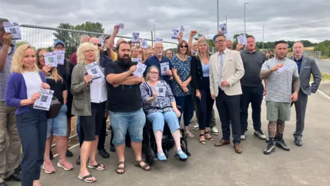 A group of people campaigning, standing outside on the pavement in front of a metal fence. Many of them are holding leaflets.