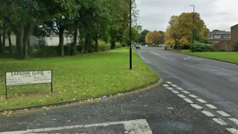 A sign for Earsdon Close is at the front of the image standing on a green lawn. It marks a junction with a much wider road, with trees and houses on either side 