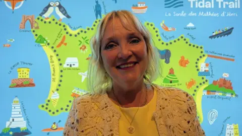 Tricia Warwick is pictured wearing a yellow dress and she has blonde hair. She is pictured in front of a map of Jersey that says tidal trail.