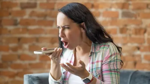 A woman with long black hair shouts into a phone she is holding in front of her. She is wearing a pink and green chequered shirt over a white t-shirt. Behind her is a brick wall.
