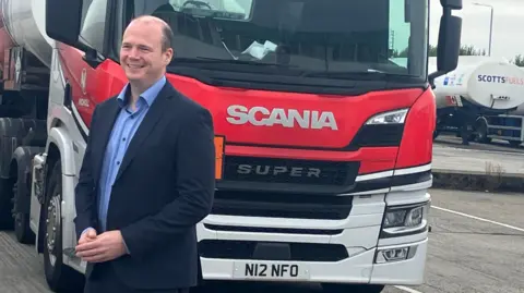 Communities Minister Gordon Lyons, wearing a blue shirt and navy suit, standing in front of a red and white truck. 