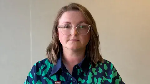 A woman with shoulder-length hair and clear glasses looks directly at the camera. She is wearing a blue and green patterned top.