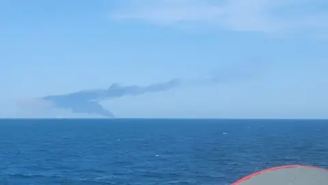Sailor provided to BBC News Black smoke rises in the distance over the horizon of the sea. A grey barrel that is part of a ship is in the right-hand corner of the image. It is a bright day with deep blue sea and a light blue sky.
