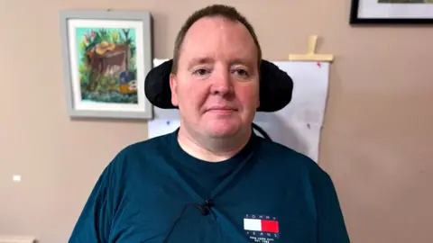 a man looking at the camera - he is in front of a wall with art hanging on it, and a wheelchair headrest is visible behind his head