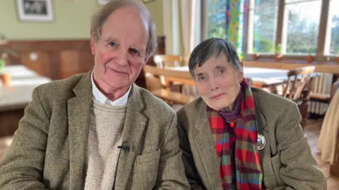 The picture shows two people seated indoors in a warmly lit room with wooden panelling along the walls. They are wearing tweed jackets and sitting at a wooden table or set of chairs. 