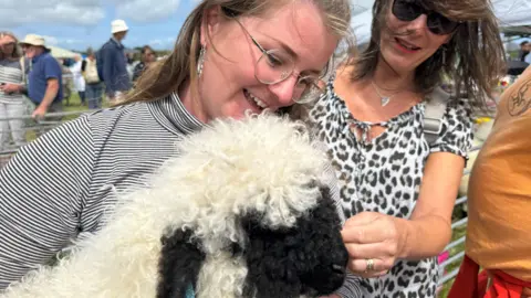 Woman in stripy top and glasses holding a lamb with a woman wearing sunglasses and a leopard print dress stroking the lamb