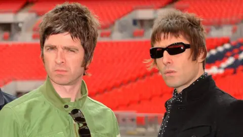 Noel Gallagher (left) wearing a green jacket with sunglasses tucked into his collar, stands next to his brother Liam Gallagher, wearing sunglasses, a black jacket and a polka dot shirt, at Wembley Stadium