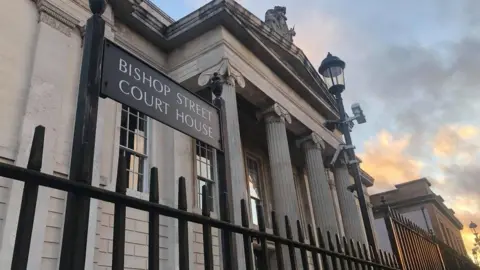 A sign for Bishop Street courthouse looms over black iron railings. Behind the railing the front of Derry's courthouse can be seen, with a lampost in front of four large pillars.