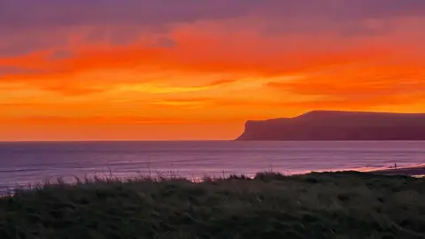 Weather Watchers/Superted A bright red and orange sunset over Marske-by-the-Sea, Redcar and Cleveland. In the foreground there is a grassy cliff edge and silhouetted against the sky there is a cliff stretching out into the sea.