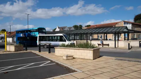 Two buses, one a small white vehicle and the other a blue double-decker, wait outside a large bus shelter with a black metal frame and glass roof. The station features stone walls, planters and pavements. A passenger stands beside a bench.