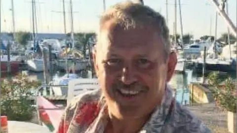Mike O'Leary wears a tropical shirt, smiling in the sunshine at a harbour, boats visible in the background