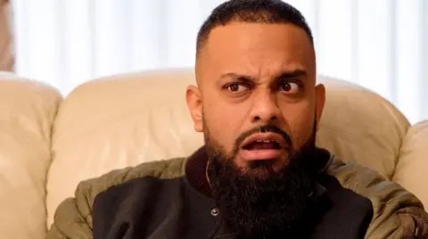 A man with short dark hair and a beard sat on a beige sofa.