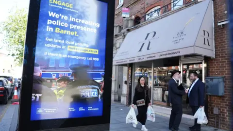 PA Media A streetside advertising display promotes increased police presence in Barnet, standing beside a shopfront on a busy urban high street. Several pedestrians carrying shopping bags walk past the storefront, while parked vehicles and brick buildings line the street in the background.