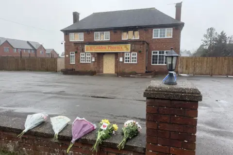 A fire-damaged building with Georgian-style windows and a sign which says "Golden Phoenix" above the door. In the foreground are five bunches of flowers, which have been placed on a wall.