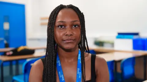 Martin Giles/BBC A 16-year-old girl with black braided hair is looking at the camera. She is wearing a black dress and has a blue school lanyard