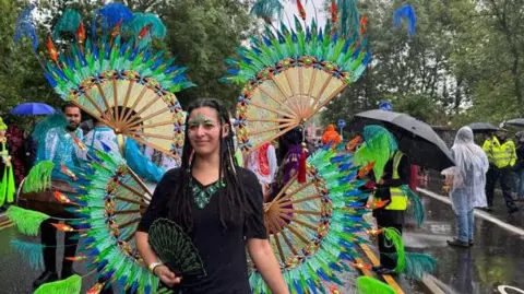 A woman wearing bright green and blue feathered wings which radiate in circles from her and shoulders. She is also carrying a fan and her face is decorated with jewels and face-paint. Her long black hair is braided and she is walking down the centre of a rainy street, with others processing behind her and onlookers lining the pavements.