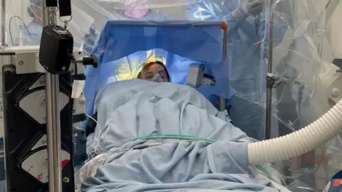 Sophie Reid Sophie lies on an operating table in a brightly lit surgical theatre, covered in blue drapes with their feet visible at the end. Several medical staff in scrubs and masks stand around the patient, with plastic drapes and equipment surrounding the operating area. Overhead surgical lights and monitoring machines are visible.