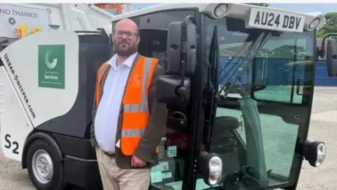 Great Yarmouth Borough Council Paul Wells wears black-rimmed glasses and has a beard. He is wearing an orange hi-vis gilet over a jacket, a white shirt and cream chinos. He is standing in front of a small street-sweeper vehicle.