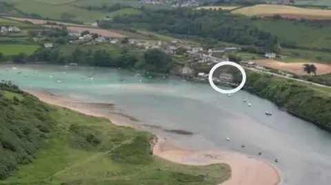 Circled in white is a boathouse with a meandering river in front, flanked on both sides by banks covered with vegetation, and on one side by residential buildings and other structures.