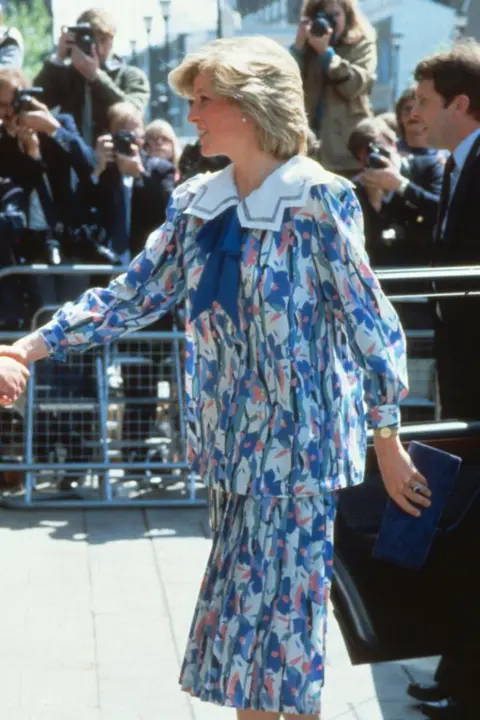Getty Images Diana, Princess of Wales, wearing a Liberty print maternity suit designed by Jan Van Velden, while pregnant with Prince Harry, visits the Albany Centre on May 23, 1984 in Deptford, United Kingdom.
It is a pastel floral outfit in blue with bits of pink, a large Peter Pan collar and a blue ribbon at the neck.