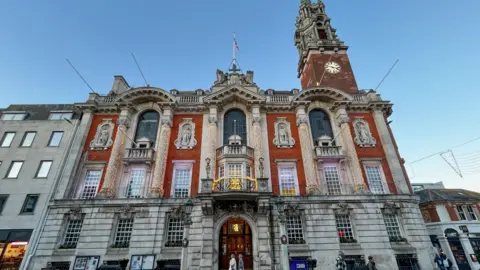 Colchester City Council A large ornate building made of grey stone with red brick sections and statues embedded into the walls. There is a large door at the front and a balcony above it. There are a number of flag poles suspended from balconies