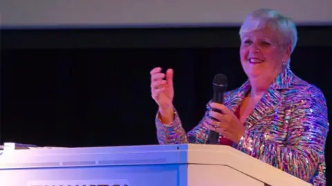 Jean Martyn A woman in a sparkly multi-coloured blazer holds a microphone up as she smiles while sitting at a white mini piano. She has short blonde hair. A black backdrop is behind her.