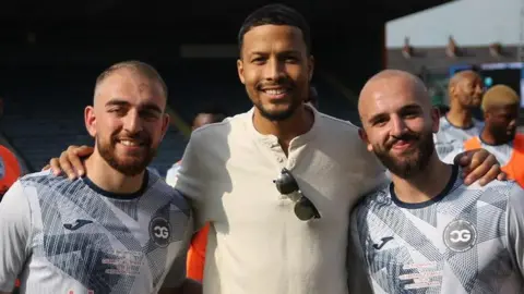 Bader Al-Akkad Amer Al-Akkad, Joe Thompson and Bader Al-Akkad stand arm in arm at a football ground, with players visible behind them.