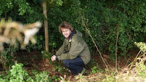 BBC/Wall to Wall Media/Stephen Hart A person in an olive-green outdoor jacket crouches beside a wire fence at the edge of dense woodland. The ground is covered with leaves, grass and low vegetation, while thick green foliage fills the background. Softly blurred plants frame the foreground, drawing attention to the secluded countryside setting.
