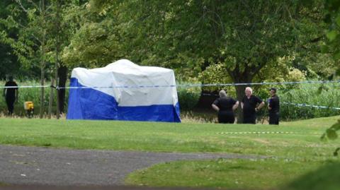 Three people in black uniforms standing by a white and blue forensics tent on the grass, which is surrounded by police tape