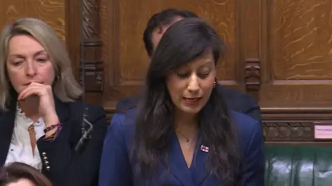Parliament TV Kumar is wearing a navy jacket and a necklace and has long dark hair.= and make up on. She is speaking in the House of Commons and other MPs can be seen behind her, a male and a female.