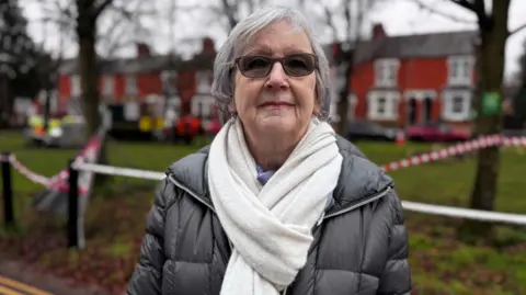 An older woman wearing a white scarf, a grey coat and black sunglasses. Behind her are trees wrapped in red and white tape.