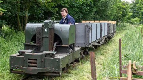 Dave Enefer/LCLR A man sat in a Simplex diesel train with two Class D bogie wagons being carried behind on a train track, surrounded by grass and trees