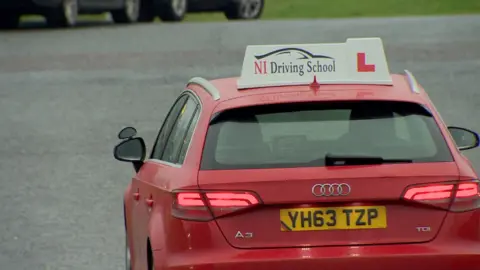 A red car with a sign on top which says 'NI Driving School' and an L sign