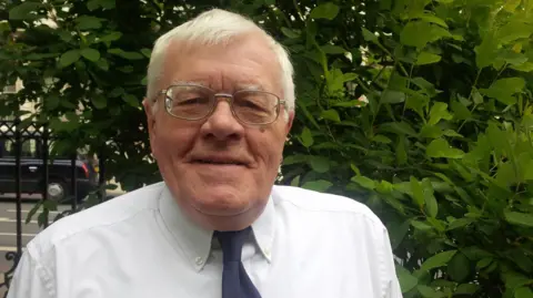 Former Lib Dem Dacorum Council leader Ron Tindall in a white shirt with a blue tie, standing by some railings and a hedge.