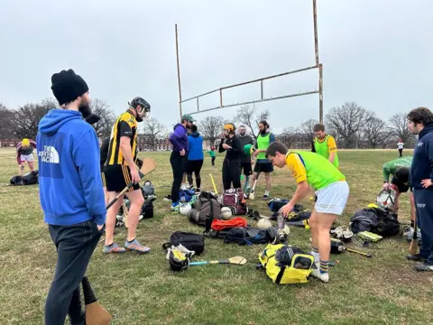 Crowd of GAA players, in sports kit, holding hurling sticks and other equipment, on a grass field