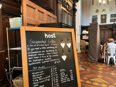 A black chalkboard sign with three hearts hanging off it and a list of drinks and their prices. The top of the sign reads: Suspended Coffee, a little act of kindness by one... to another. 
