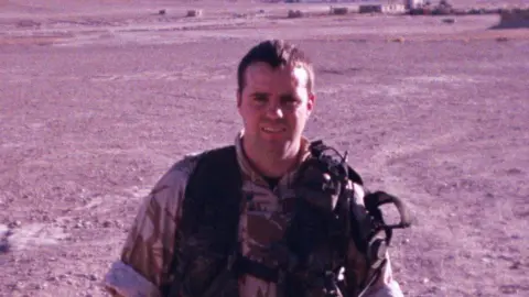 Kevin Russell looks directly at the camera standing in a desert. He is wearing Army combat gear. He has brown hair. 