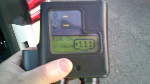 Jersey Police A close-up view shows a handheld electronic device encased in a stitched black leather cover, being held over grey tarmac. The device features a small digital display with a greenish background and black text, showing numerical readings including “+111” prominently, along with smaller figures such as “0100.22n” and a time reading “17:59:24.” Above the screen are two buttons labeled “OFF” and “TEST.” Two cylindrical attachments extend from the bottom of the device. In the background, part of a red and white object, a white vehicle car partially visible along the left side of the frame.
