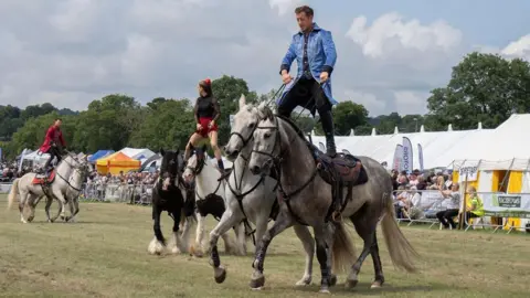 BBC Weather Watchers/Videoman A man in a blue coat and black trousers stands with a foot on the back of two horses. Behind him, a woman in a black and red outfit does the same on two different horses. Behind them, an audience looks on, in front of white tents.