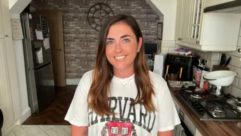 A woman in her thirties with long brown hair smiling at the camera in a kitchen. She is wearing a white t-shirt that says "harvard" on it.