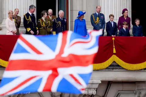Pool via Getty Images The Duchess of Edinburgh, the Duke of Edinburgh, Vice Admiral Sir Tim Laurence, the Princess Royal, King Charles III, the Duke of Kent, Queen Camilla, the Prince of Wales, Prince George, the Princess of Wales, Prince Louis and Princess Charlotte all stand on the balcony in formal dress while the King and Queen stand on the balcony of Buckingham Palace with a union flag in the foreground.