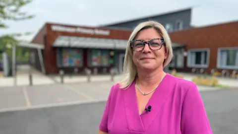 Head teacher Hazel Richards stands in front of Wilkinson Primary School, she's wearing a pink dress, has blonde hair and wears glasses. She is smiling at the camera. The school is a modern building with brown tiles on it.
